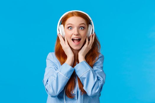 Excited and amused attractive caucasian redhead woman in pyjama, screaming from fascination and amazement hear new song, wearing headphones enjoy music, standing blue background