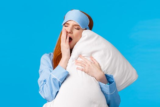 Feminine and glamour lovely redhead teenager yawning and cover opened mouth, feeling sleepy and tired got to bed, hugging pillow, close eyes having good night, standing blue background