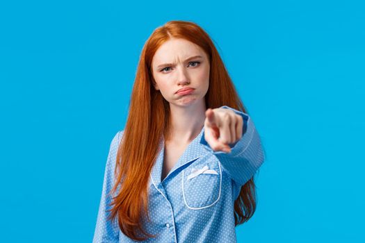 Offended timid and silly redhead teenager, girl in nightwear pointing at camera with blame, shame someone, sulking and frowning angry, accuse person, standing blue background unsatisfied