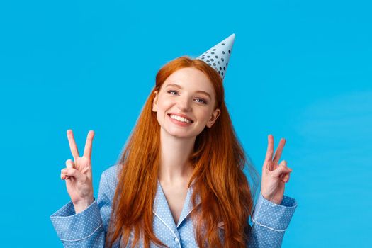 Happy cheerful redhead woman in birthday cap, tilt head and smiling upbeat, celebrating b-day, showing peace sign delighted, having sleepover party with friends, blue background