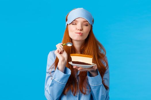 Delicious food, lifestyle and people concept. Delighted and happy cute redhead female in pyjama and sleep mask, close eyes and licking lips as eating tasty cake, holding spoon smiling