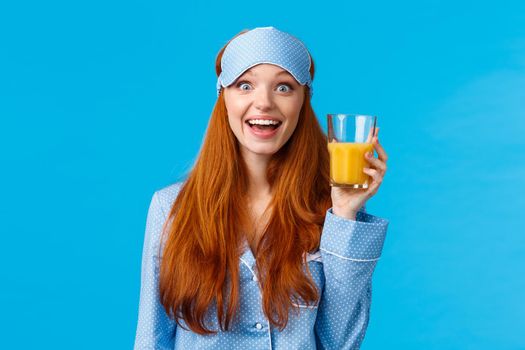 Breakfast, healthy lifestyle and women concept. Attractive smiling, excited redhead female in sleep mask, looking amused with wondered expression, holding glass of orange juice, blue background