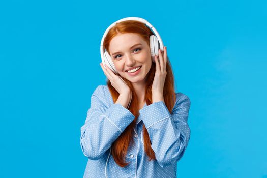 Technology, lifestyle and people concept. Cute redhead woman in pyjama, listen music in headphones, enjoy nice beats in earphones, good quality, new songs, standing blue background