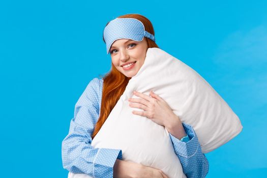 Sleepy time. Lovely and feminine glamour redhead girl with sleep mask, wearing nightwear, hugging pillow and smiling, wishing sweet dreams, standing blue background upbeat