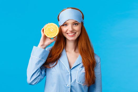 Start your morning right. Diet, healthy lifestyle and beauty concept. Cheerful glamour redhead woman in nightwear, holding slice orange near face, smiling, wearing sleep mask and pyjama