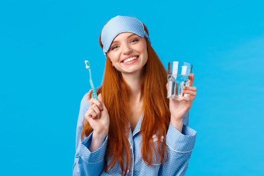 Happy smiling and carefree redhead enthusiastic girl in sleep mask, pyjama, holding glass water and toothbrush, taking care mouth hygiene, dental daily routine, blue background