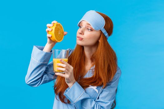 Silly and cute redhead caucasian girl in sleep mask and nightwear, looking with hopeful and eager expression, like eating fruits, squeeze orange in glass to make breakfast juice, blue background