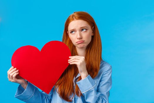 Gloomy girl was rejected on valentines day. Uneasy upset redhead female in nightwear, holding heart card and sobbing, look away upset, feeling depressed, standing blue background