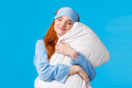 Beauty, tenderness concept. Happy feminine cute redhead teenage girl in nightwear, sleep mask, close eyes and smiling delighted, finally went bed after long day, hugging pillow, blue background
