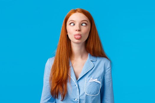 Be funny and silly. Redhead carefree and enthusiastic caucasian girl squinting, making goofy expression, showing tongue, standing blue background in nightwear, fool around