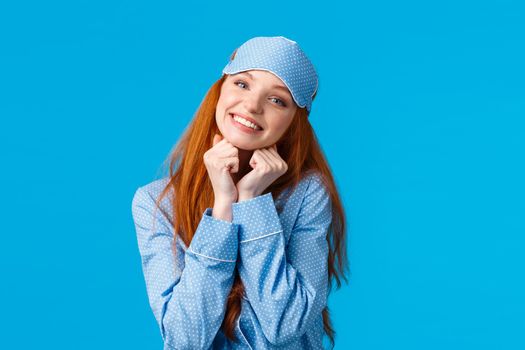 Tender, feminine redhead woman with toothy beautiful smile, tilt head holding arms near face lovely pose, smiling as standing in nightwear and sleep mask, ready have sweet dreams, blue background