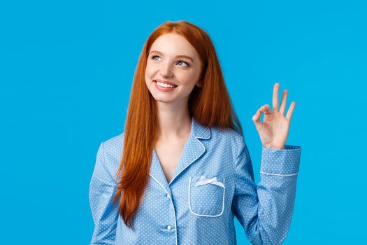 Dreamy and creative cute redhead woman feeling happy and relieved, wearing nightwear, showing recommend, okay gesture, approving new hygiene product, standing blue background
