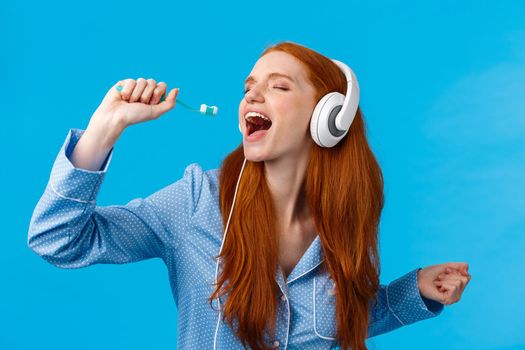 Passionate and carefree cute foxy european redhead woman in pyjama, close eyes wearing headphones, listening music singing into toothbrush, brushing teeth and getting ready morning, blue background