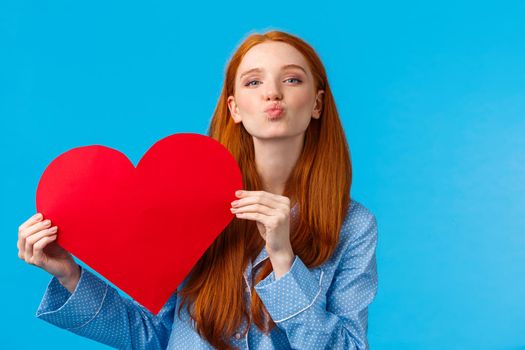 Tenderness, relationship and valentines day concept. Caring and romantic cute foxy teenage girlfriend holding red heart card and folding lips, blowing kiss, give mwah, blue background