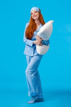 Full-length vertical studio cheerful lovely caucasian woman with ginger red long hair in nightwear, pyjama and sleep mask, hugging pillow ready sleep going bed at nigh and laughing, smiling