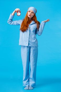 Why didnt you set alarm. Confused and unsure upset attractive redhead woman in pyjama and sleep mask, shrugging perplexed staring questioned camera, holding clokc, blue background