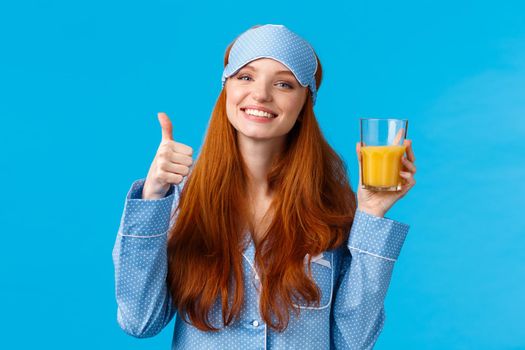 Drinking fresh juice is healthy and good. Cute glamour redhead caucasian female in sleep mask and nightwear, tilt head and smiling holding drink in glass and show thumb-up smiling