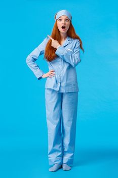 Full-length vertical shot excited and intrigued cheerful redhead female in nightwear and sleep mask, open mouth saying wow, have you seen it, pointing upper left corner amazed, blue background