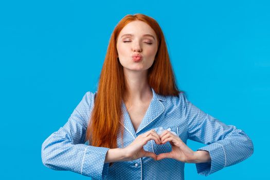 Kiss for goodnight. Charming pretty redhead female in pyjama folding lips and close eyes give mwah, showing heart gesture over chest, express affection or tender feelings, blue background
