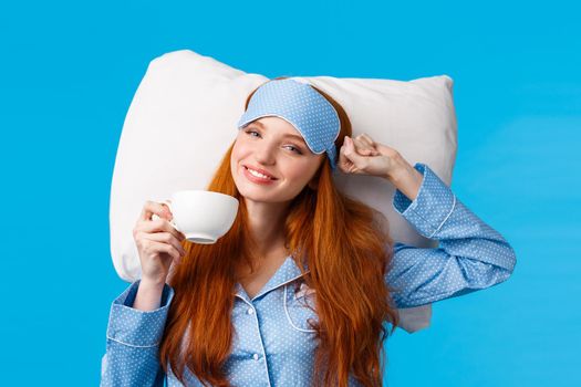 Perfect morning of princess. Cheerful, tender redhead woman with long ginger hair, wearing sleep mask and pyjama, stretching in bed feeling fresh and energized, drink morning coffee, blue background