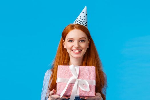 Waist-up portrait cheerful happy, pretty redhead, ginger girl celebrating birthday, receive cute pink wrapped present, wearing b-day cap and smiling, having fun at party, blue background