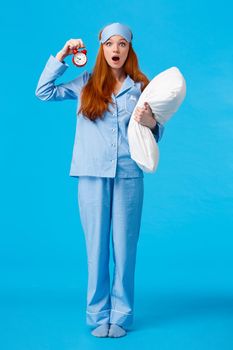 Vertical full-length shot shocked and startled cute redhead female in sleeping mask and pyjama, holding pillow and alarm clock, open mouth gasping worried, stare camera, blue background