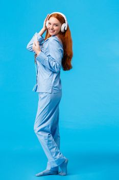 Vertical full-length portrait sassy and coquettish, flirty redhead woman in cute pyjama, dancing listen music in big white headphones, hold smartphone, using app to enjoy favorite playlist