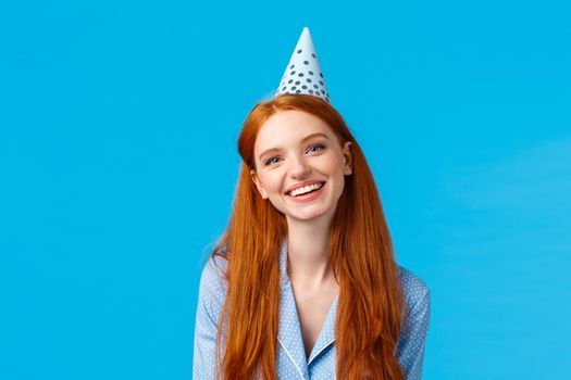 Happiness, beauty and celebration concept. Cheerful carefree redhead woman in b-day hat and nightwear, smiling joyfully, have sleepover birthday party, standing blue background