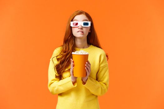 Lifestyle, hobby and people concept. Cute geeky redhead woman in 3d paper glasses, holding popcorn and biting lip as staring at favorite actor playing in movie, visit cinema to watch premiere