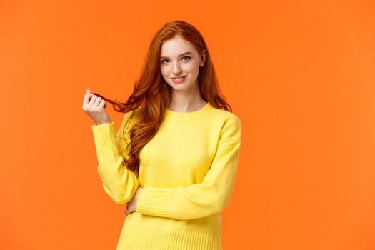 Gorgeous flirty and cute young modern redhead cheeky girl, rolling curl on hair and glancing camera curious, with interest and coquettish flirting smile, standing orange background intrigued