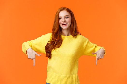 Waist-up portrait cheerful redhead female student in yellow sweater, inviting check-out event, recommend proudct, chrismas sale, holiday promo, pointing down and smiling, orange background