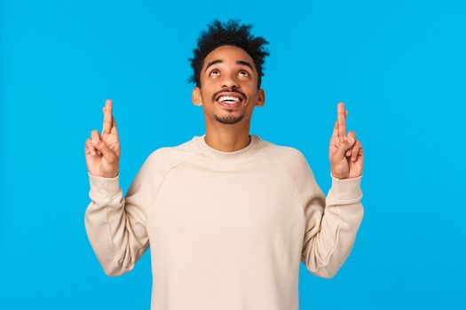 Enthusiastic and intrigued modern african-american male in white sweater, seeing something curious upwards, look and pointing fingers up, smiling astonished, wonder whats there, blue background