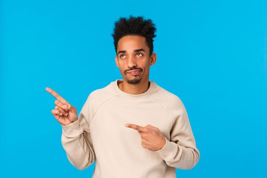 Skeptical and disappointed handsome stylish african-american modern guy in white sweater, smirk and staring with judgement and dislike at something lame, pointing upper left corner, blue background