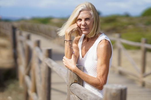 Happy female in her 60s leaning on wooden fences on the sand of a tropical beach.