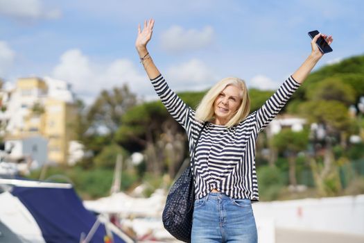 Woman in her 60s strolling along a seaside spot near the beach, raising her arms in joy.