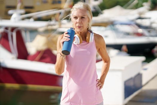 Mature sportswoman taking a break during exercise to hydrate herself. Senior woman in fitness clothing drinking water from a metal fitness bottle. Concept of healthy living in the elderly.
