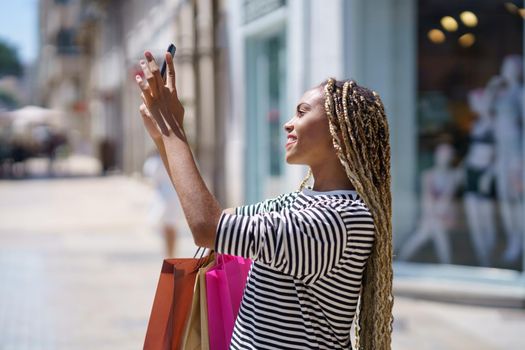 Young black woman photographing something in a shopping street with her smartphone.