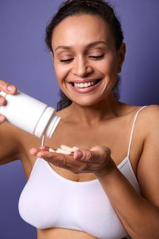 Smiling cheerful African American woman in white underwear holds a bottle of food supplement and pours a vitamin complex into her hand. Medical pills for healthy skin. Health care and medicine concept