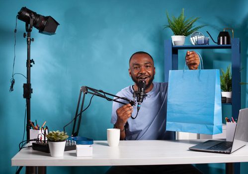 Vlogger holding boom arm microphone presenting giveaway blue paper gift bag in vlogging studio