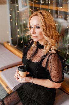 Young charming blonde with a cute smile and makeup while relaxing in a cafe. She is holding a cup of coffee in her hands. She is dressed in a black dress with transparent sleeves.