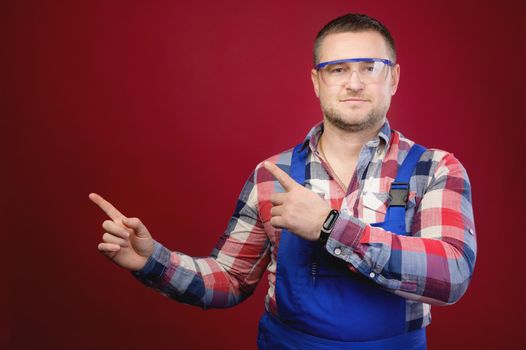 Attractive Caucasian male foreman in uniform and goggles points fingers at blank space. Foreman repairman maste studio portrait
