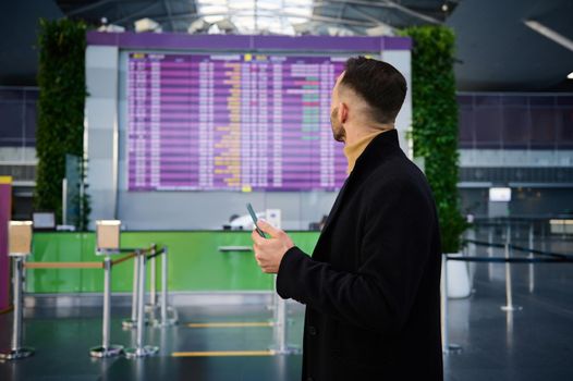 Handsome young man, businessman on a business trip, dressed in a dark coat, with a phone in his hands, looks at the information panel of the timetable at the international airport