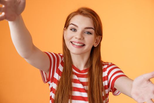 Cheerful friendly redhead 20s woman extend arms, hold camera both hands taking selfie, tilt head cute, record video blog new gadget, rejoice stand orange background, show lovely grin