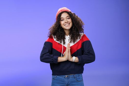 Portrait of charming curly-haired feminine girlfriend begging for favour as posing over blue background in winter outfit with hat, holding hands in pray smiling with angel look and friendly gaze