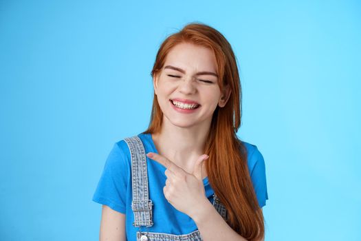 Cheerful carefree redhead woman sincere smile, pointing left introduce copy space joyfully close eyes, show white toothy grin delighted, feel upbeat rejoicing, joyful positive mood, blue background