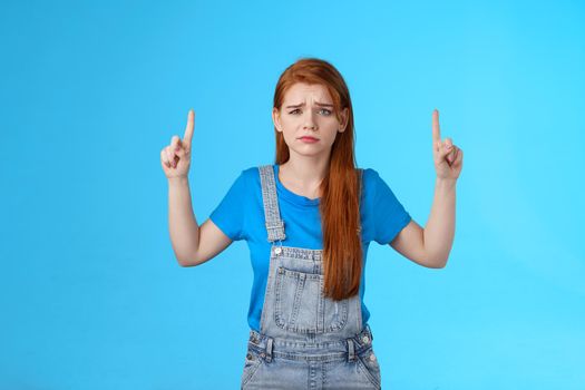 Doubtful concerned unsure redhead female making important decision, grimacing hesitant displeased, point up promo, top copy space, feel uncertain unlikely buy suspicious product, blue background