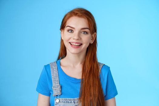 Friendly confident pleasant redhead woman helpfully look camera, smiling joyfully, have conversation customer, give advice, stand upbeat good positive mood blue background, grin amused