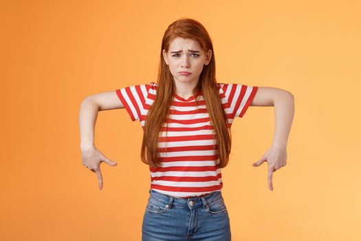 Cute timid hesitant ginger female taking tough decision feel pressured upset, frowning, pulling gloomy unhappy face, pointing down disappointed, uneasy taking decision, orange background