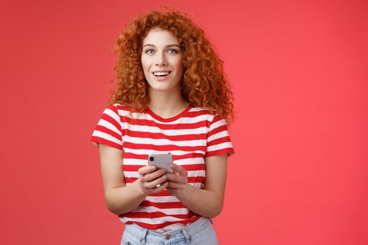 Fashionable carefree good-looking relaxed redhead girlfriend hold smartphone look camera excited pleased turn attention friend answering messages social media writing post red background