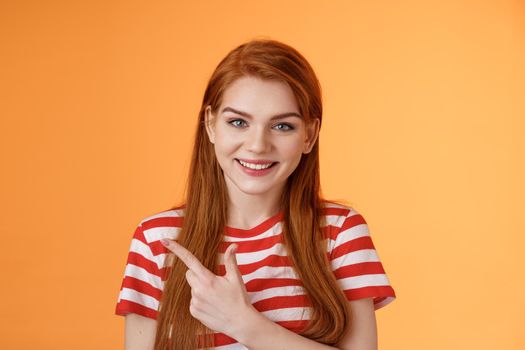 Close-up cheerful smiling girl pointing sideways. Redhead woman introduce promo gladly recommend good offer, grin discuss interesting place indicate upper left corner, orange background